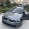 A Volkswagen Golf 6 1.6 TDI in gray, with the driver's door open, is parked on a driveway in front of a modern building adorned with trees and potted plants. The car's trunk is also open.