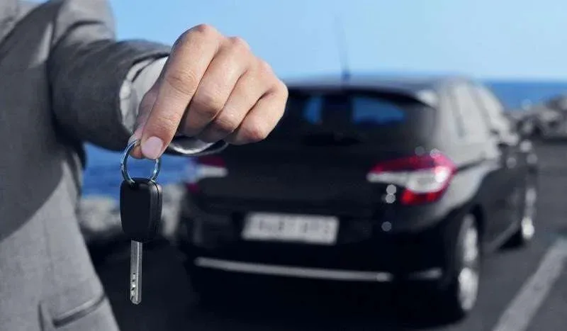 A person in a gray suit holds car keys in front of a black car parked by the sea. The background shows a clear blue sky and rocky shoreline.