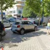 A street scene featuring a Volkswagen Polo Cross parked among other cars on a cobblestone road. A person in a yellow shirt and denim shorts walks along the sidewalk, while trees and buildings can be seen under a sunny, clear sky in the background.