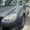 A Volkswagen Golf 5 in gray is parked on a paved street in front of a building with a HOTEL sign, showcasing a modern design with angular features. Small trees line the sidewalk.