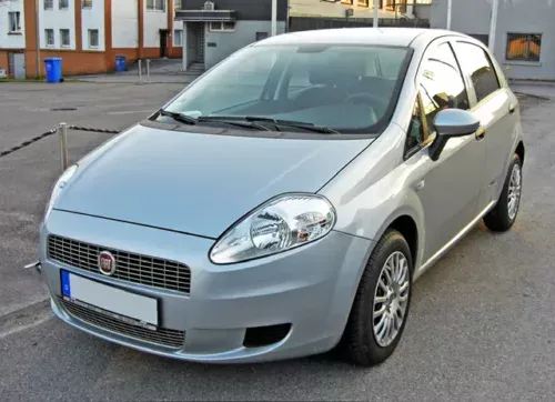 A Volkswagen Polo Cross in a silver hatchback design is parked on a street in an urban setting, showcasing its sleek design and shiny surfaces, with residential buildings and blue recycling bins visible in the background.