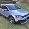 A Volkswagen Polo Cross in silver is parked on a grassy area beside a road, featuring a sleek design with tinted windows and alloy wheels. Trees and shrubs can be seen in the background.