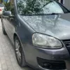 A Volkswagen Golf 5 in silver is parked next to a sidewalk on a tiled street, with a sign in the background that reads VETEM PER HOTELIN. Trees and a building can be seen behind the car.