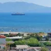 A view from the Sweetview 2-bedroom apartment with seaview on three balconies reveals houses and greenery in the foreground, a tranquil blue sea with a large ferry in the middle, and hazy mountains under a clear blue sky in the background.