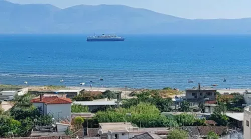 A view from the Sweetview 2-bedroom apartment with seaview on three balconies reveals houses and greenery in the foreground, a tranquil blue sea with a large ferry in the middle, and hazy mountains under a clear blue sky in the background.